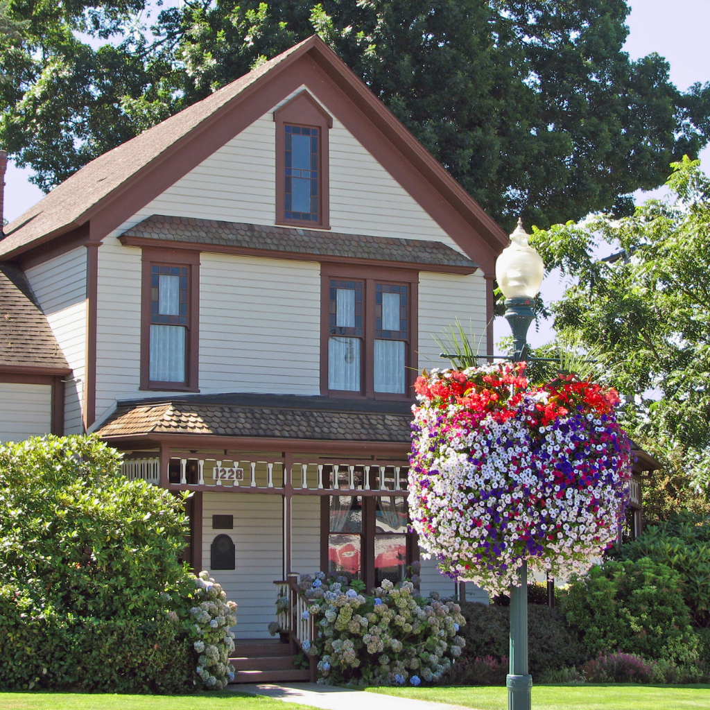 A charming two-story house with cream-colored siding and brown trim, surrounded by lush greenery and blooming flowers. A decorative streetlamp in front of the house is adorned with a large, colorful hanging basket of red, purple, and white flowers.